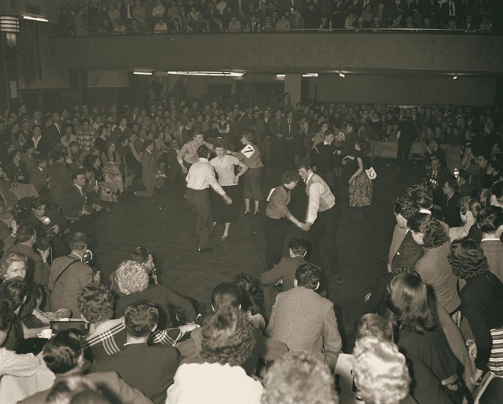 A packed hall watches young couples jive in a rock and roll dance contest.