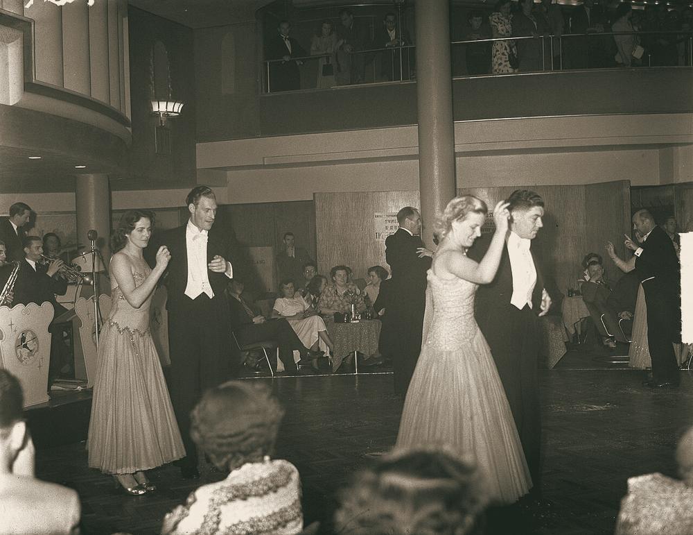 Couples in evening dress dance across a ballroom floor while people watch from tables and a balcony.