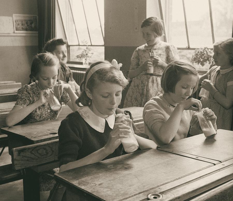 Schoolgirls at wooden desks drinking their school milk through straws.