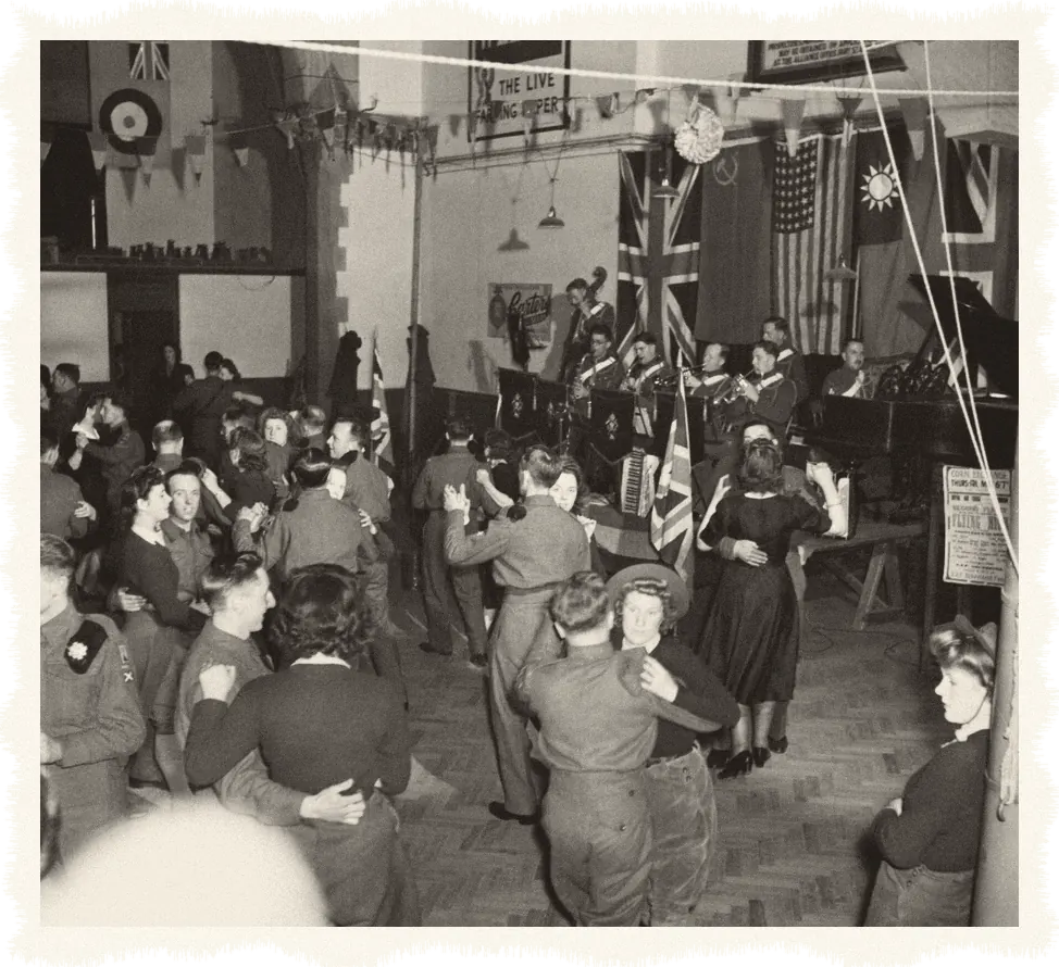 Couples dance in a crowded hall hung with flags and bunting while a band plays on the stage