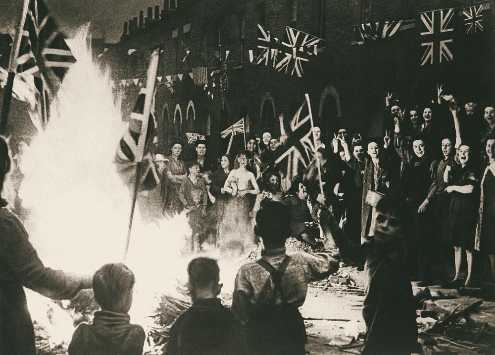 A crowd round a big street bonfire, flags strung overhead, children in front.