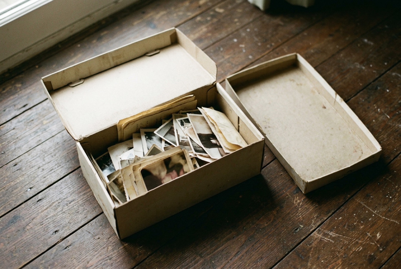 An open shoebox of loose old photographs and papers on a wooden floor