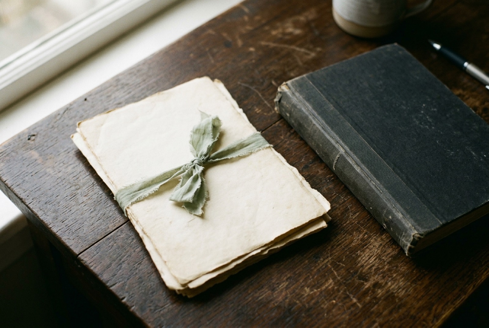 A small bundle of aged letter paper tied with a ribbon, beside a worn closed notebook on a wooden desk